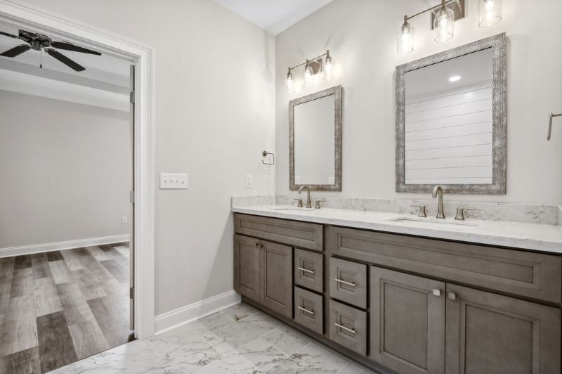 Bathroom with double vanity, gray cabinets, white countertops, and marble-like flooring. A door to bedroom is visible.