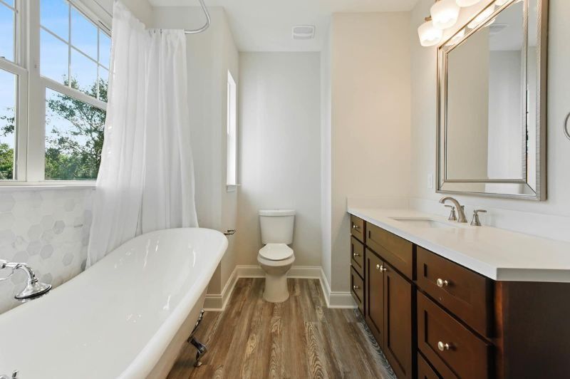 Bathroom with a white bathtub, vanity, and toilet. Light wood-look flooring and a large window.