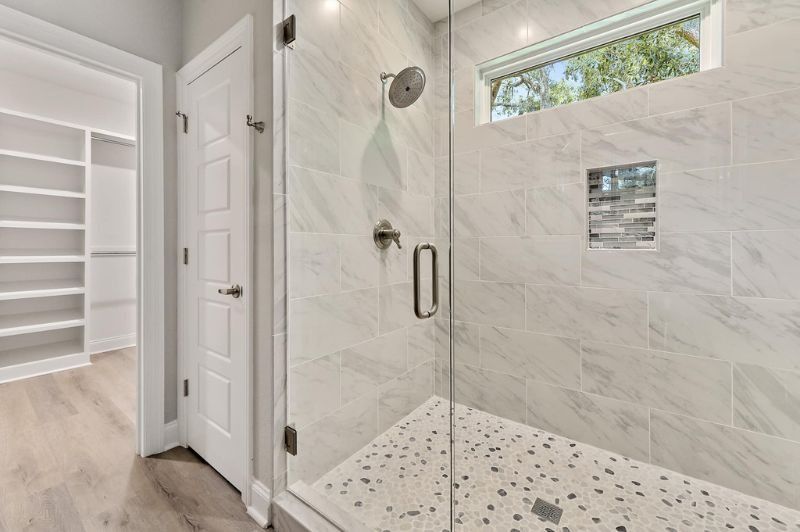 Bathroom with glass shower, marble tile walls, walk-in closet entrance, and a pebble stone shower floor.