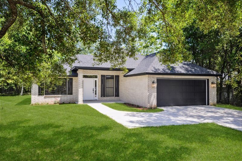 A single-story light brick house with a black garage door under trees, on a green lawn.