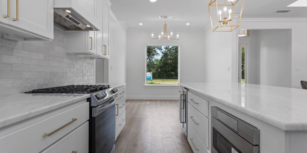 White kitchen with island, cabinets, and stove. Window with trees visible. Gold light fixtures and cabinet pulls.