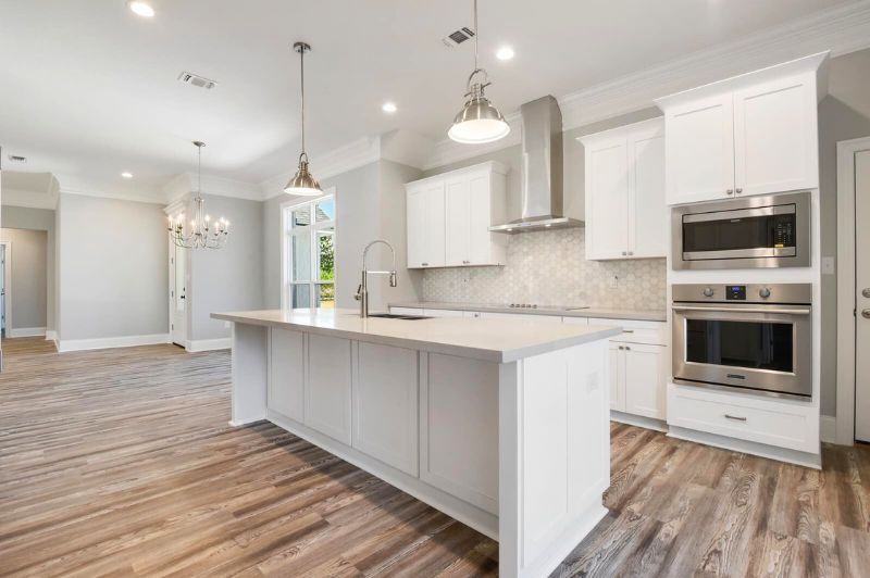 Modern white kitchen with island, stainless steel appliances, light wood floors, and pendant lights.