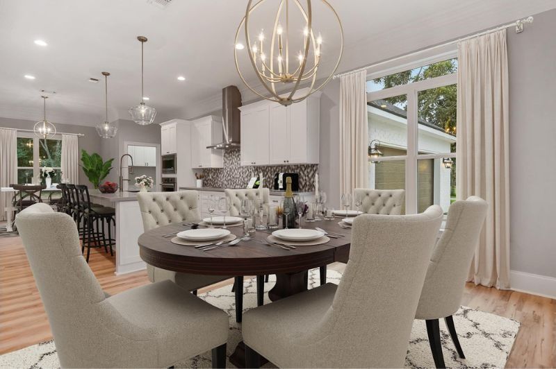 Dining room with round table, upholstered chairs, chandelier, and kitchen visible in the background.