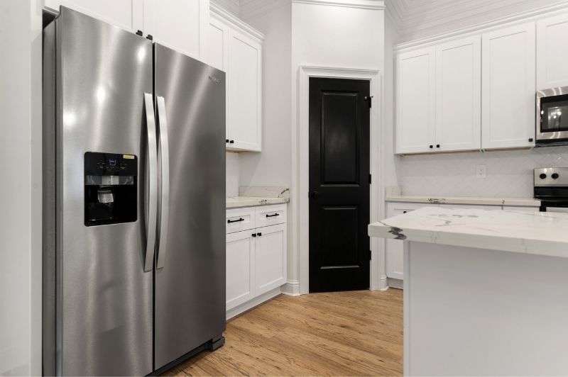 Stainless steel refrigerator in a bright white kitchen with wood floors and a black door.