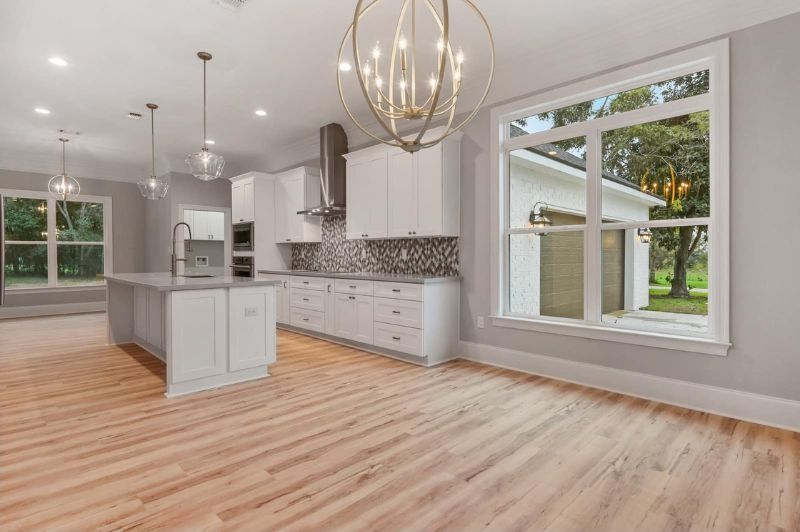 Modern kitchen with white cabinets, island, and hardwood floors, open to a window and a chandelier.