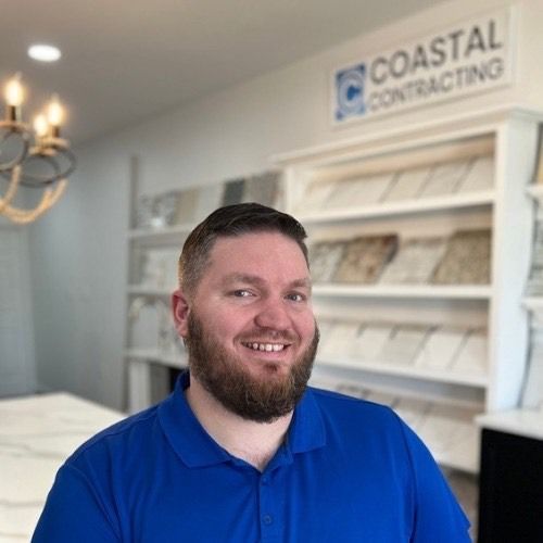 Man with a beard smiles in front of Coastal Contracting sign and tile samples. He wears a blue shirt.