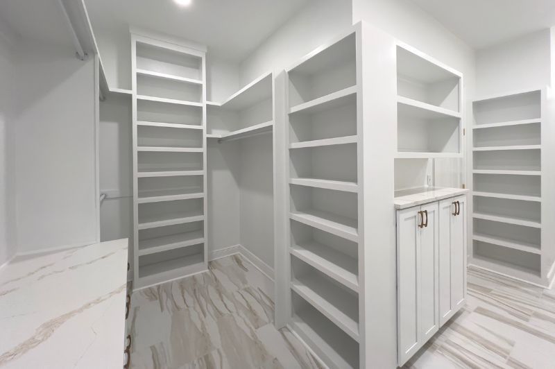 White walk-in closet with built-in shelves and cabinets, light-colored flooring.