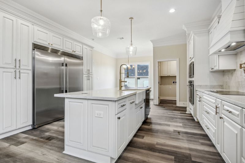 White kitchen with large island, stainless steel refrigerator, and wooden floors.