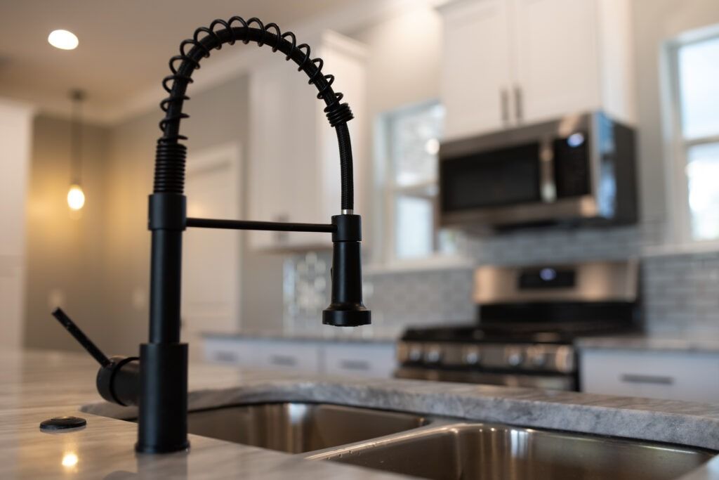 Black kitchen faucet over a stainless steel sink, blurred background of kitchen appliances.