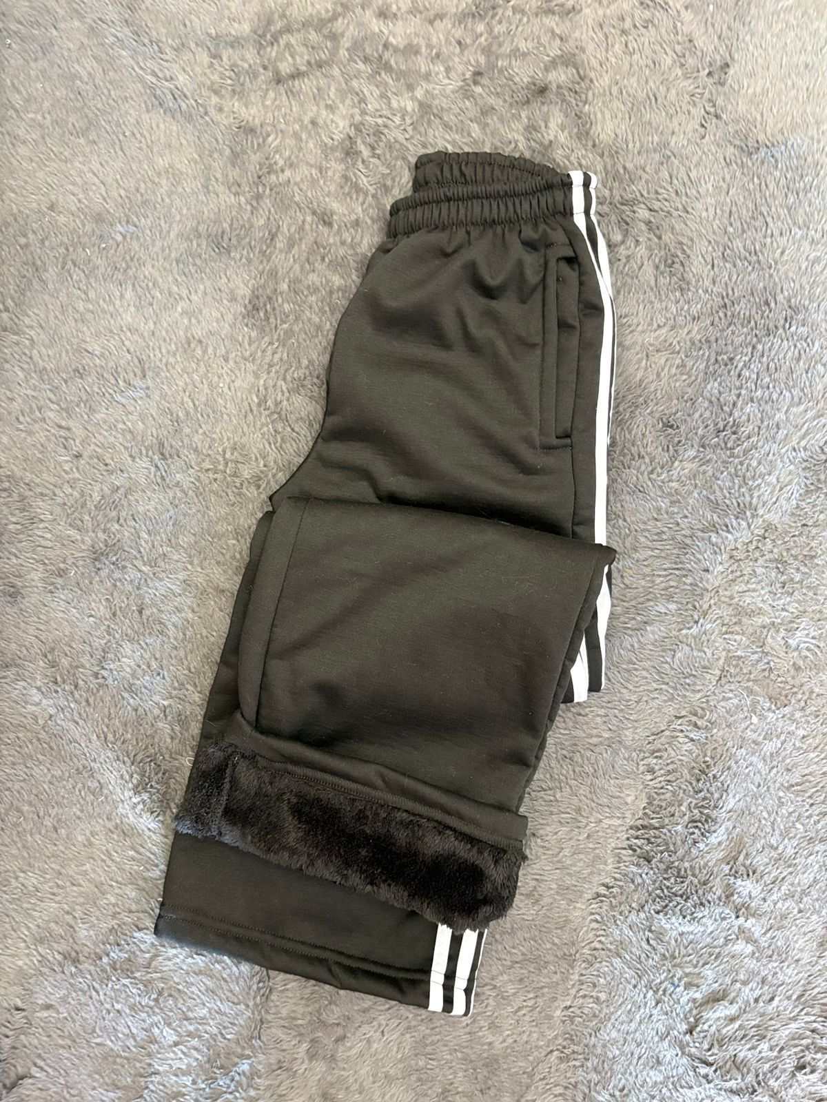 Un par de pantalones deportivos negros con rayas blancas en los laterales están sobre una alfombra.