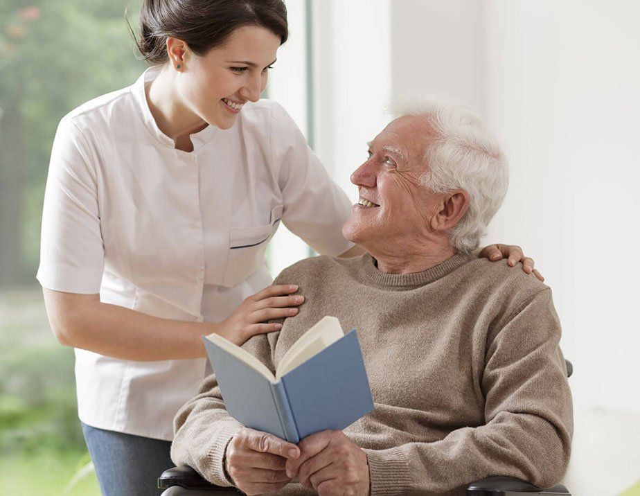 A nurse is helping an elderly man in a wheelchair read a book.