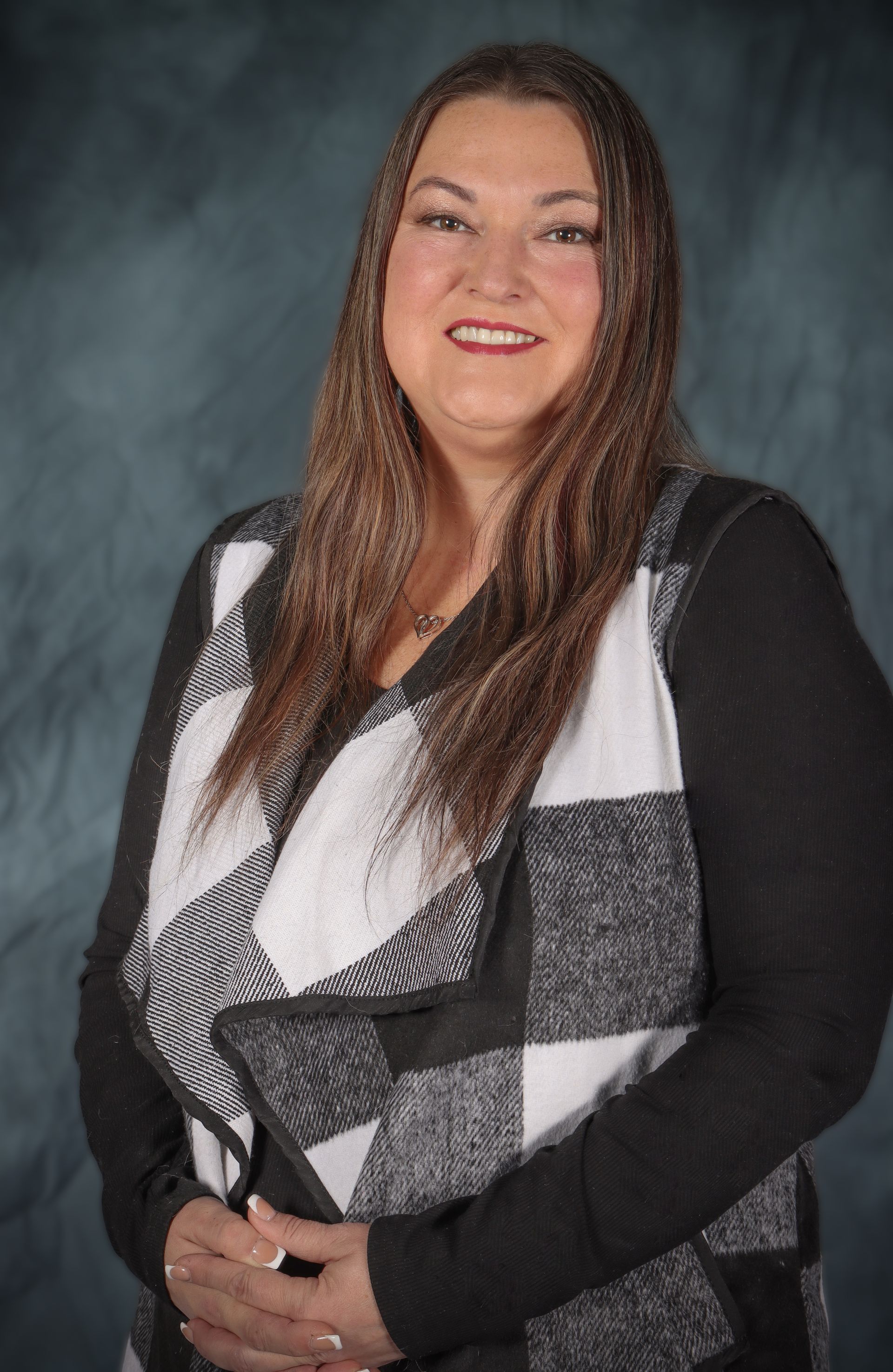 Woman with long brown hair, smiling, wearing a black and white vest over a black shirt, posing against a blue-gray backdrop.