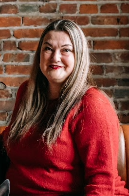 Woman with long blonde hair, red sweater smiles in front of a brick wall.
