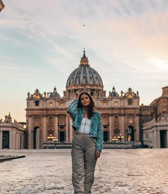 Woman in front of St. Peter's Basilica, posing. She is wearing a blue jacket, white top, and tan pants. Dusk setting.