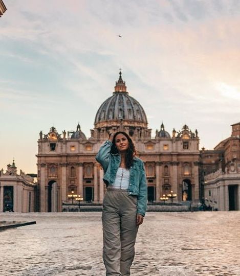 Woman in front of St. Peter's Basilica, Rome. Wearing denim jacket and pants, looking at the camera. Golden light.