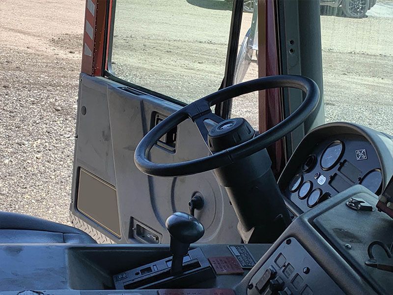 Interior of a truck cab, with steering wheel, dashboard, gear shift, and open door to the outside.