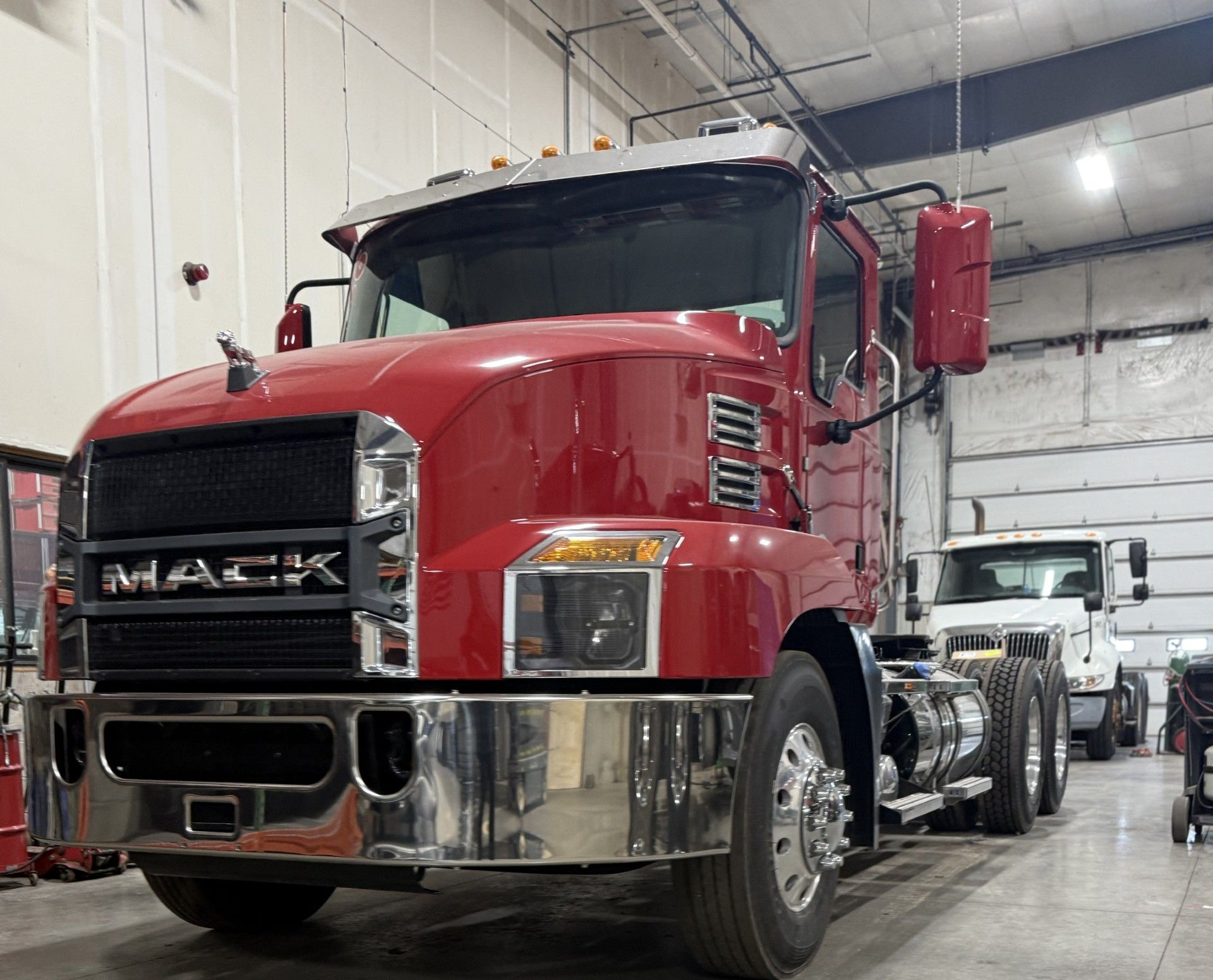 Red Mack semi-truck parked inside a warehouse. Chrome bumper, black grill, and a white truck in the background.