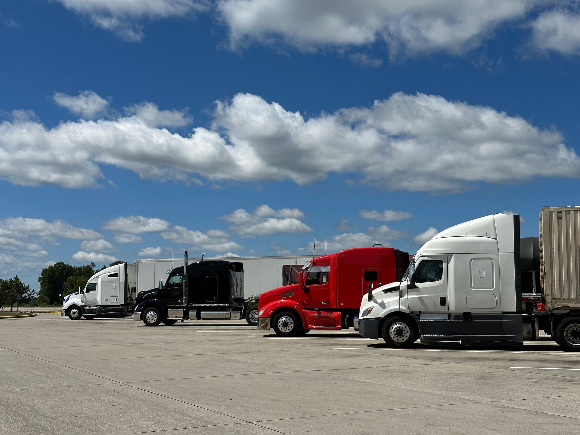 Semi-trucks parked in a lot under a blue sky with fluffy clouds.