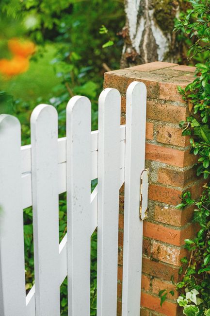 White picket gate attached to a brick pillar, green foliage in the background.