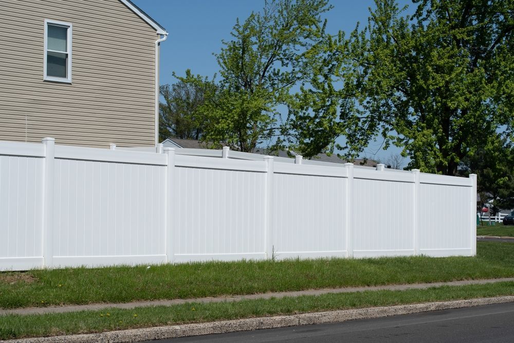 White vinyl fence in front of a house with green grass, next to a road and blue sky.