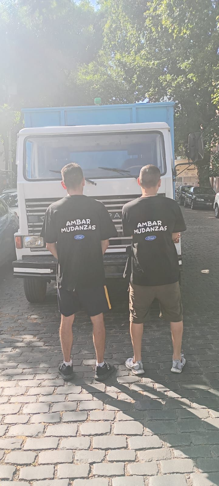 Dos hombres con camisas negras están de pie frente a una camioneta blanca, de espaldas a la cámara. 