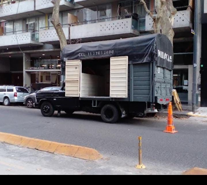 Camión negro estacionado en la calle, con cajas de madera visibles. Cono naranja y edificio al fondo.