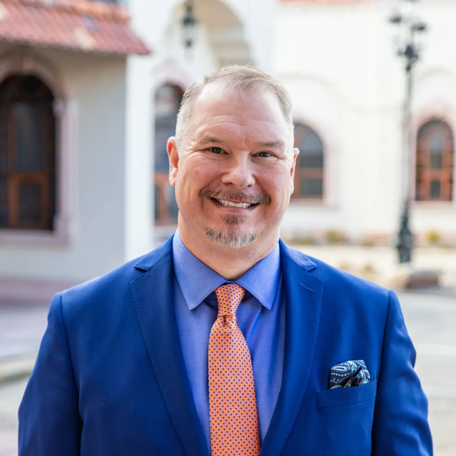 A man in a blue suit and orange tie is smiling for the camera.