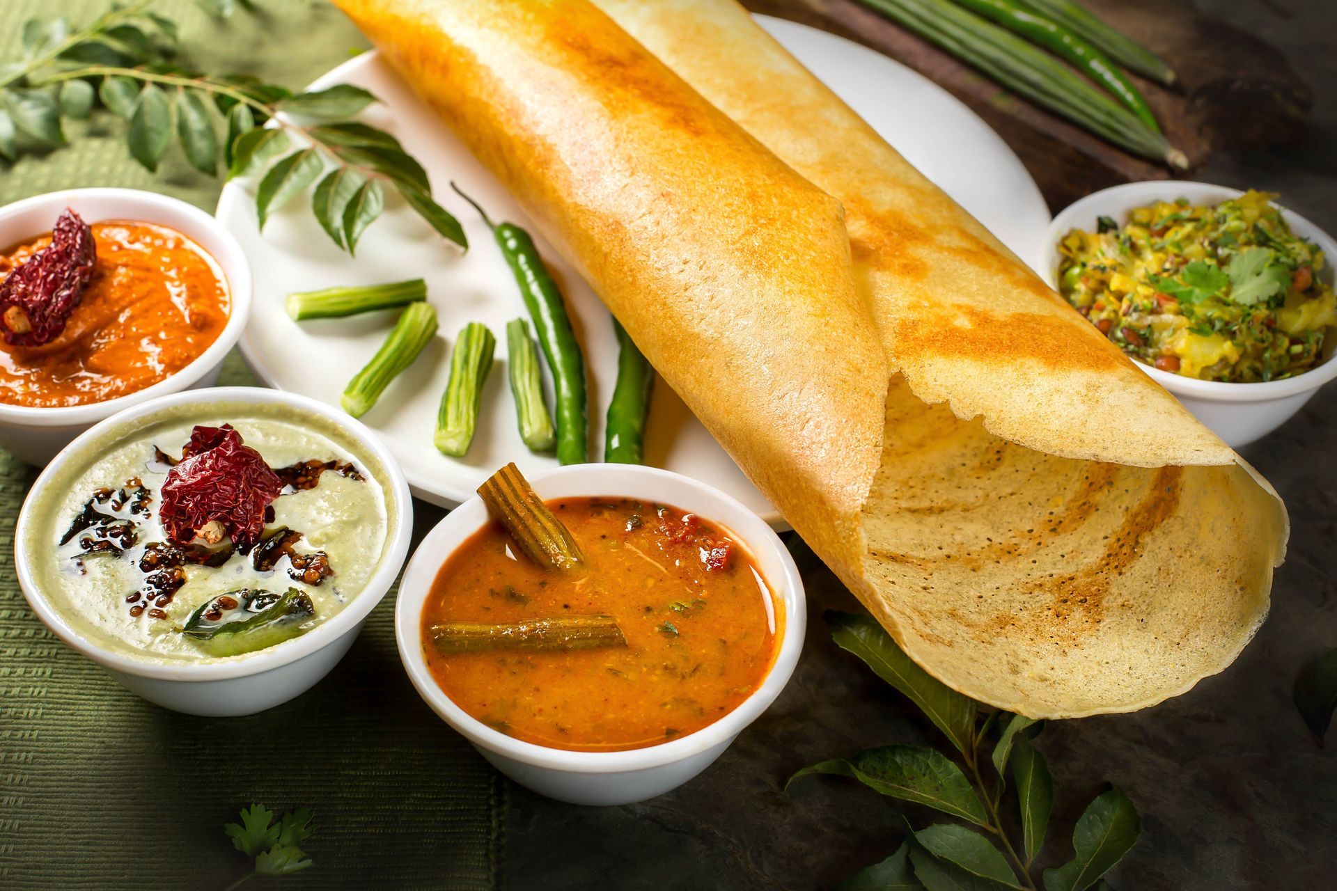 Crispy dosa with various chutneys and sambar, green chilies on a white plate.