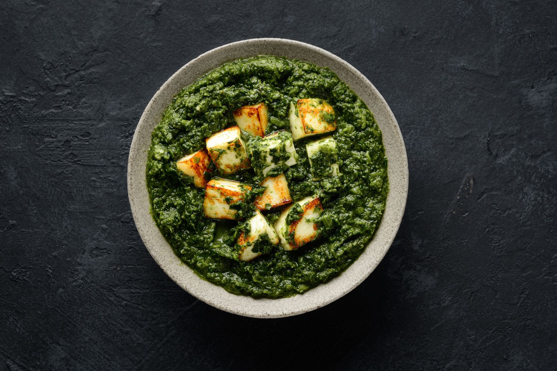 Palak paneer: Cubed paneer cheese in a green spinach-based curry, served in a grey bowl on a dark surface.