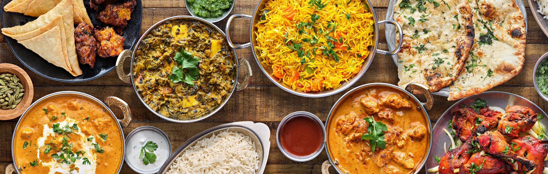 Overhead view of Indian food spread on wooden table: curries, rice, naan, and sides.