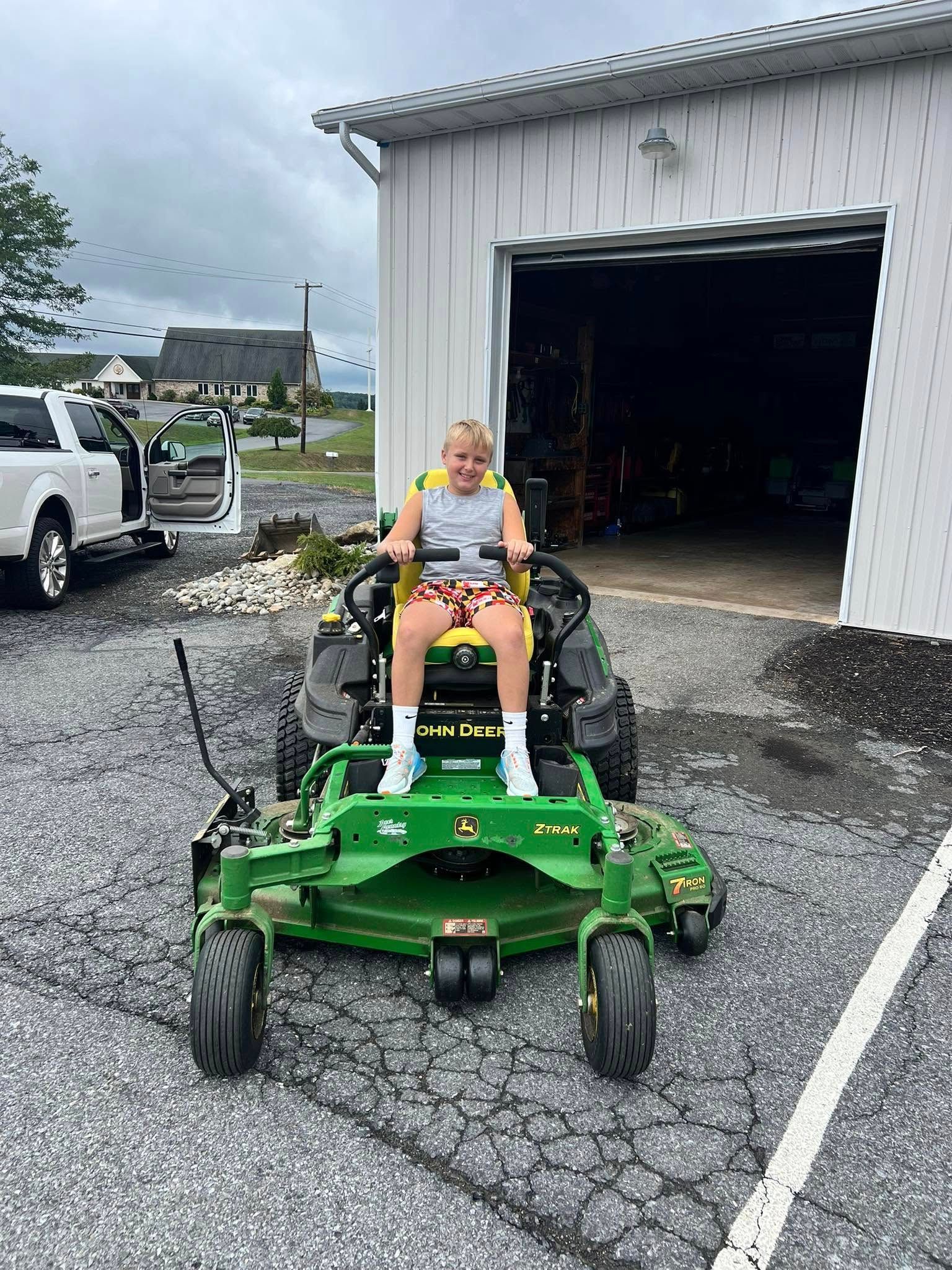 Child sitting on a John Deere riding lawnmower in front of a garage, smiles.