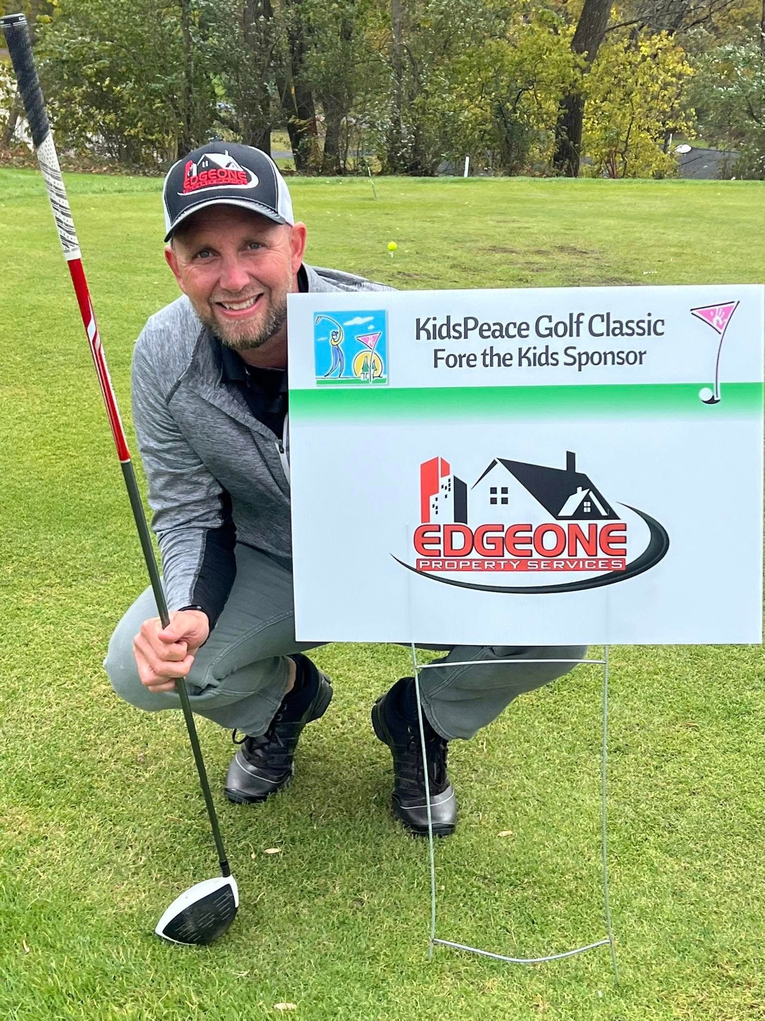 Man kneels on a golf course, holding a golf club and sign. He's smiling. The sign is a Kids Power Golf Classic sponsor.