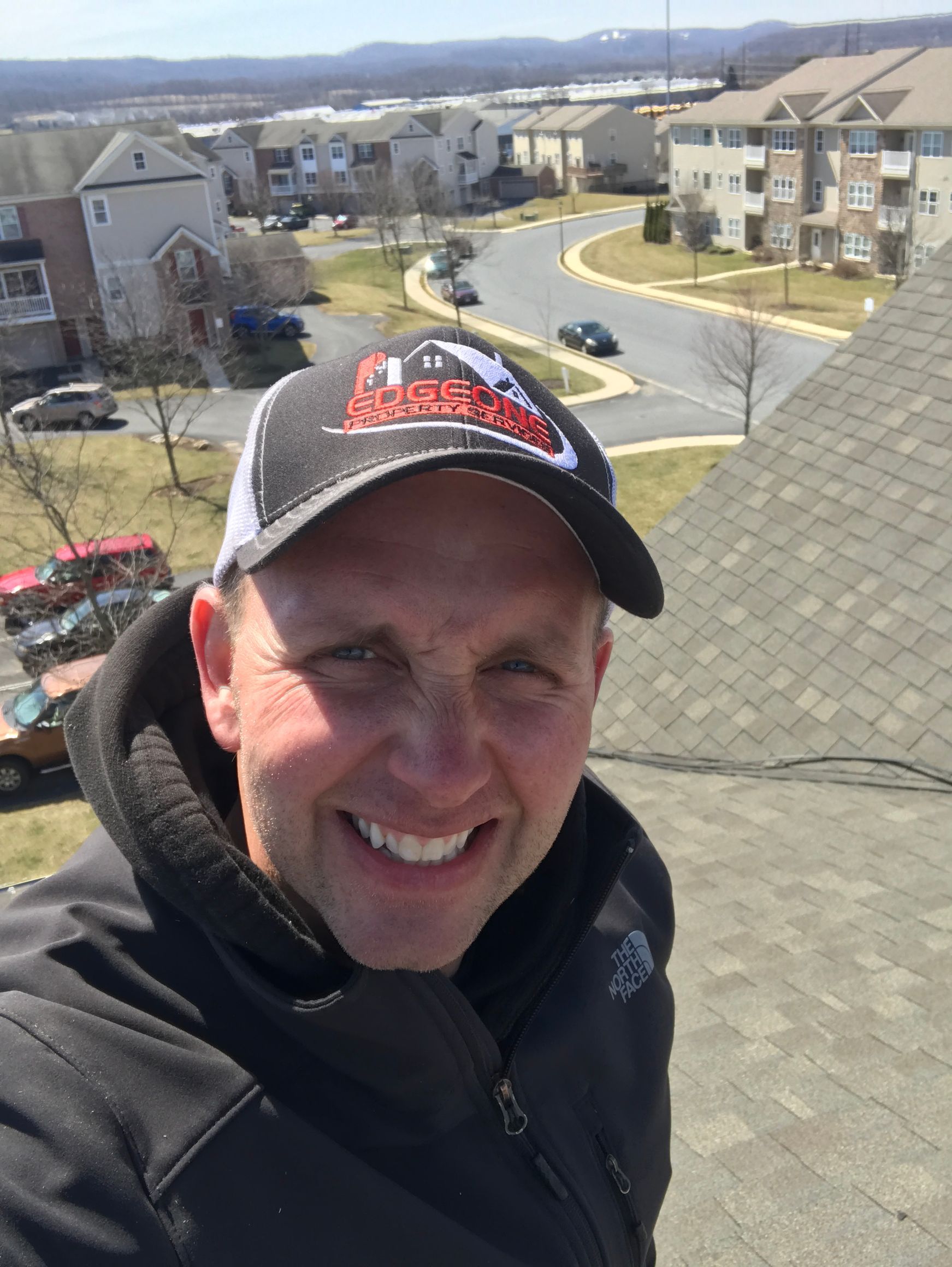 Man on a rooftop wearing a cap, smiling, with a suburban neighborhood in the background.