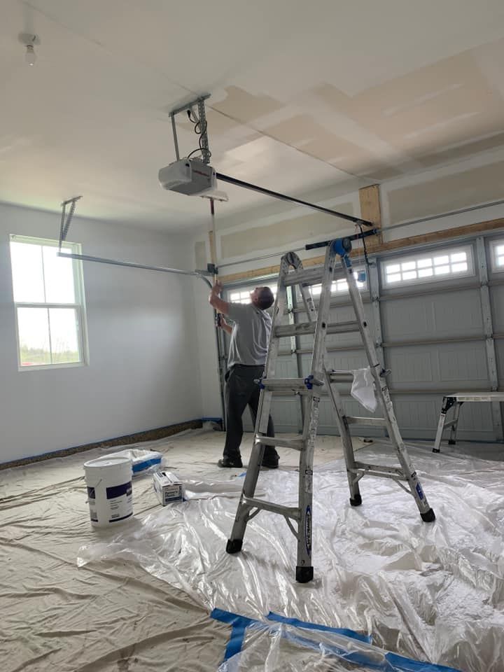 A person on a stepladder works on a garage door opener. White walls, a window, and paint cans are in the garage.
