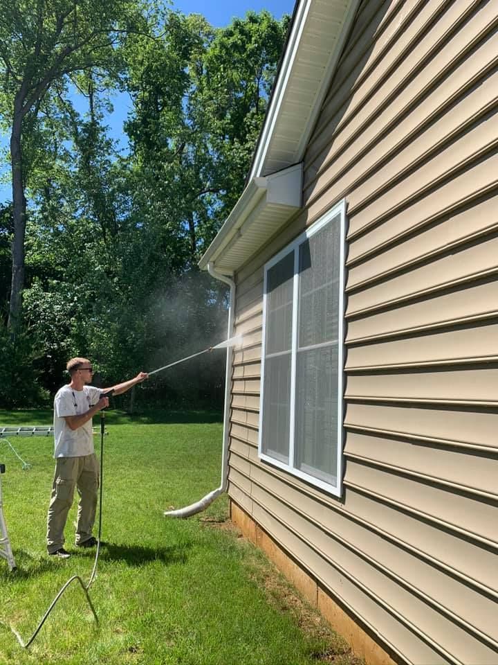 Person power washing siding on a beige house with white trim. Green yard, sunny day.