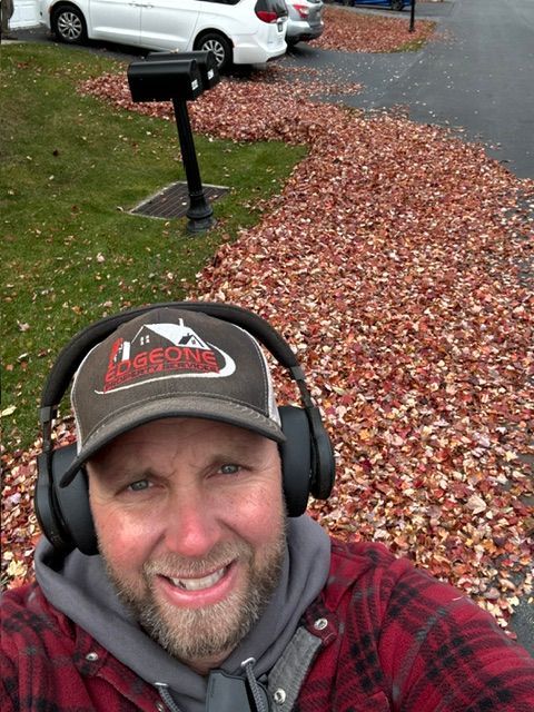 Man with earmuffs smiles, surrounded by fall leaves, near a mailbox and cars.