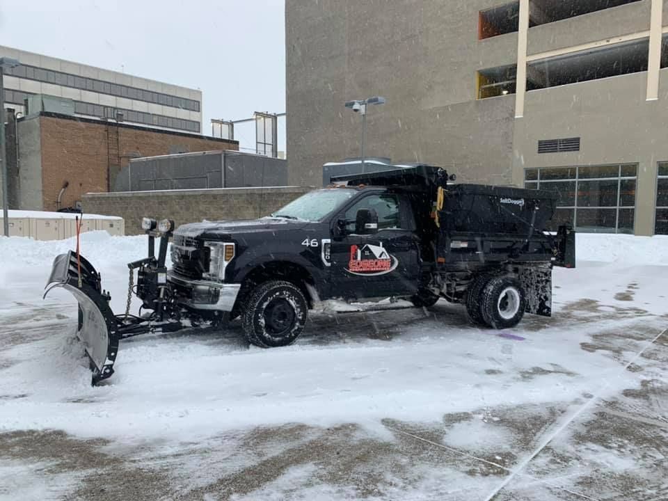 Black snow plow truck plowing snow in a parking lot; snowy day.