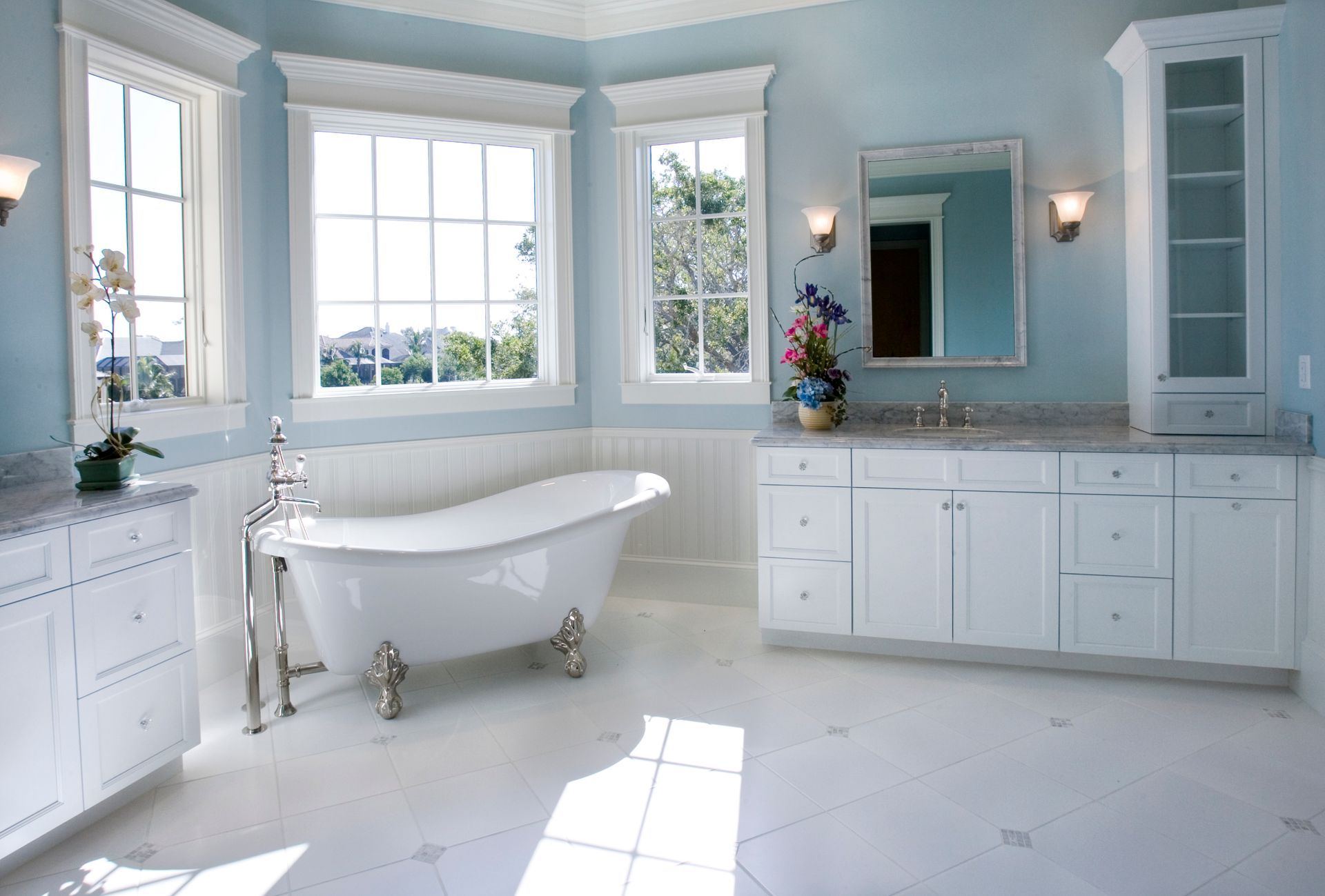 Bright blue bathroom with white cabinets, tub, and large windows.