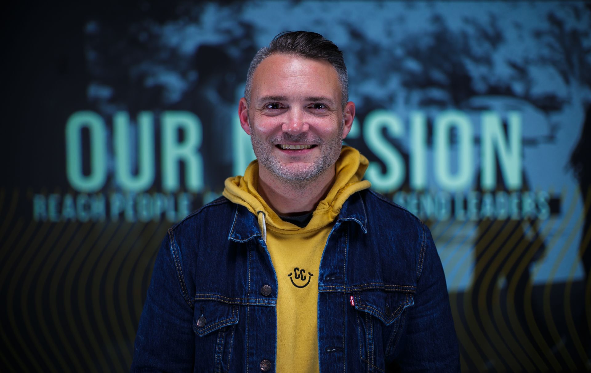 A man wearing a denim jacket and a yellow hoodie is smiling in front of a sign that says `` our mission ''.