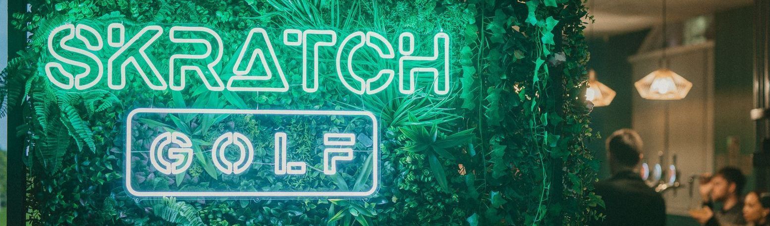 Four women on a golf course, celebrating near a hole with a flag. They are on a green lawn, with trees, a pond, and overcast skies.