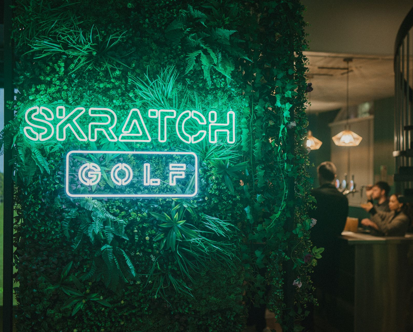 Neon SKRATCH GOLF sign on a lush green wall inside the venue