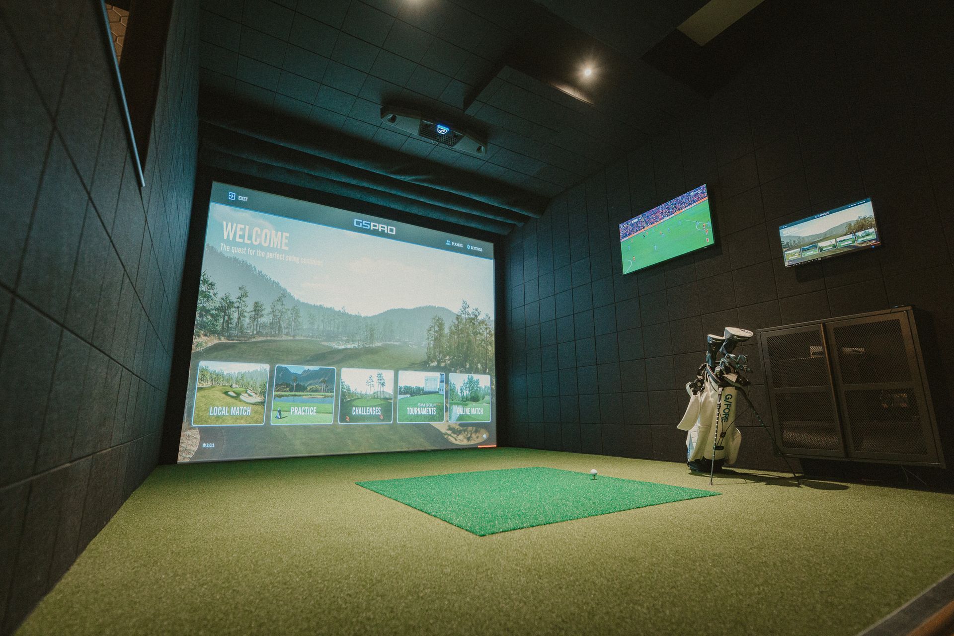 A golf simulator room with a large screen displaying a golf course, a green mat, and golf clubs on the right.