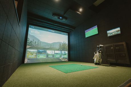 Golfer practicing indoors in a simulator bay, swinging on a mat toward a virtual fairway screen.