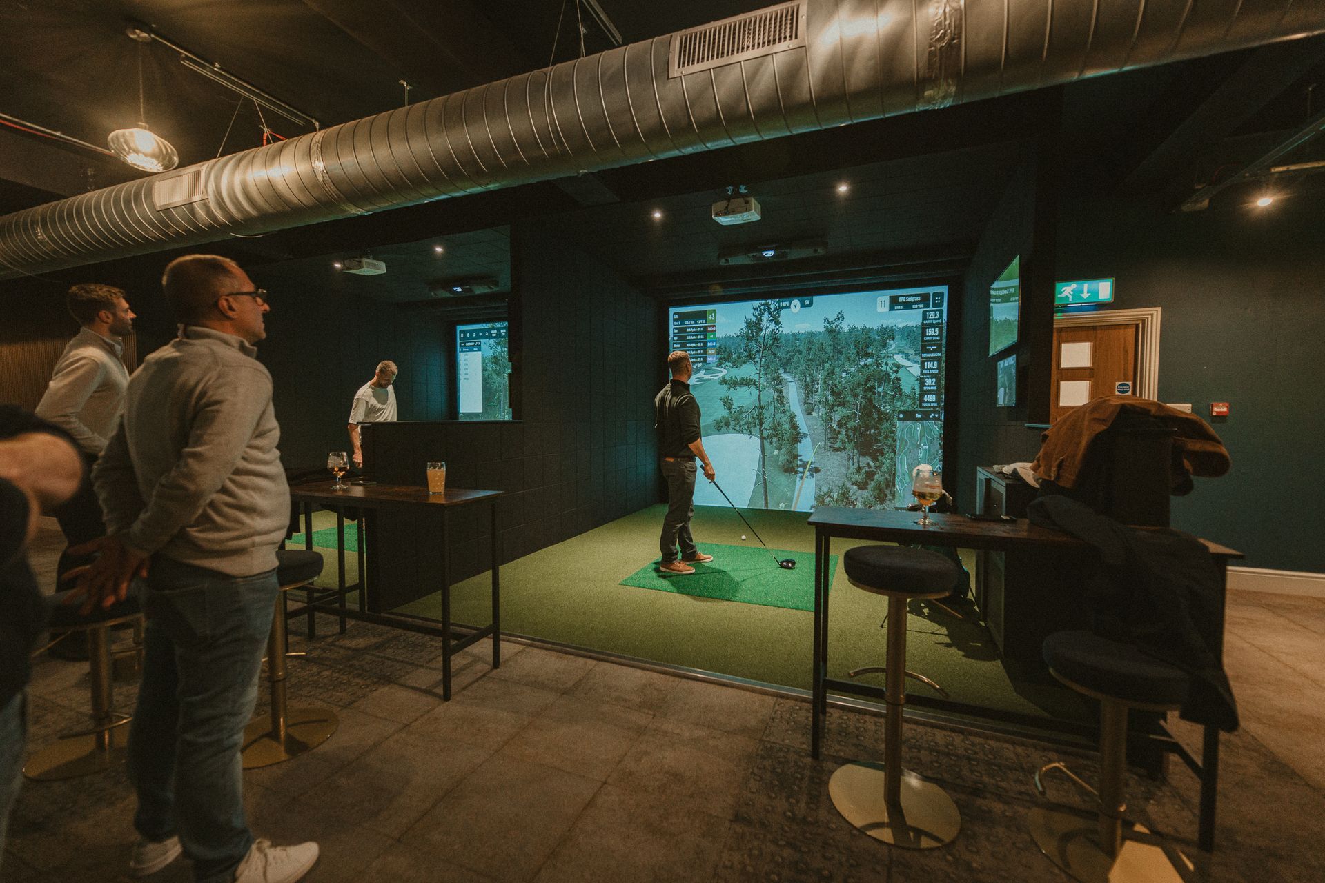 Indoor golf simulator with people observing a golfer. Dark setting with screens, bar tables, and stools.