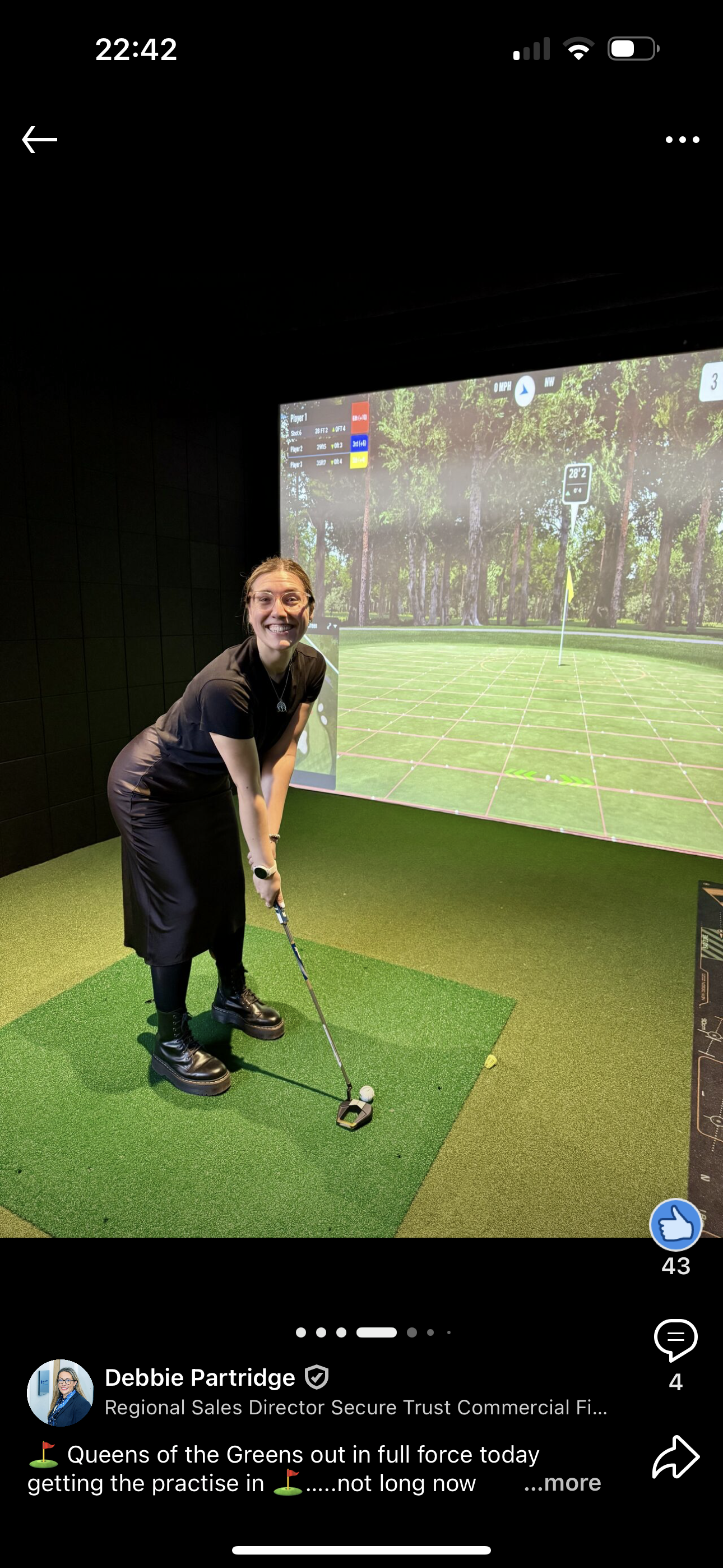 Woman in black clothing smiles while practicing golf indoors. She is on a green mat with a screen showing a golf course in the background.