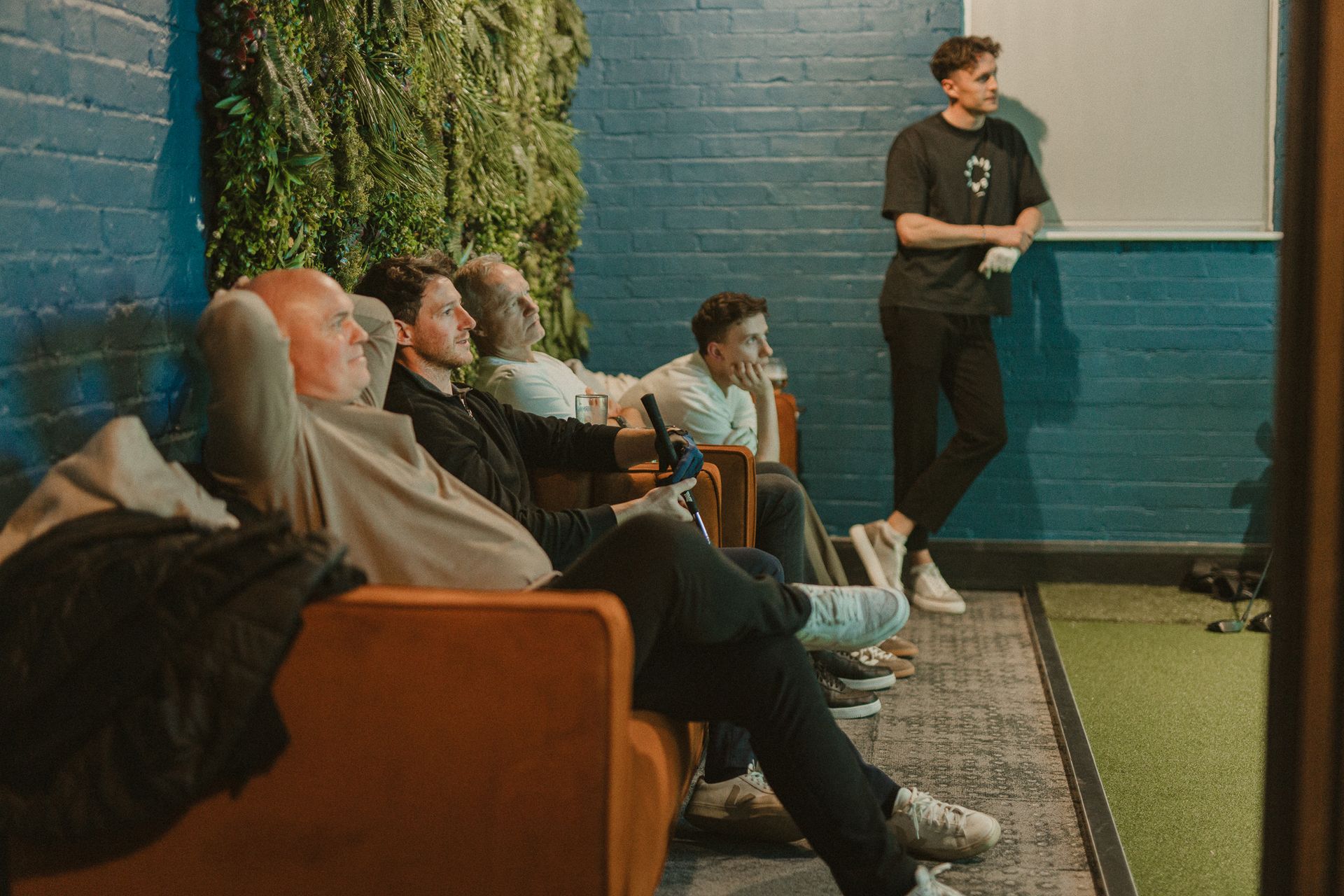 People in a lounge-like space; some on a couch, another leaning near a screen. A wall has vertical greenery and dark blue paint.