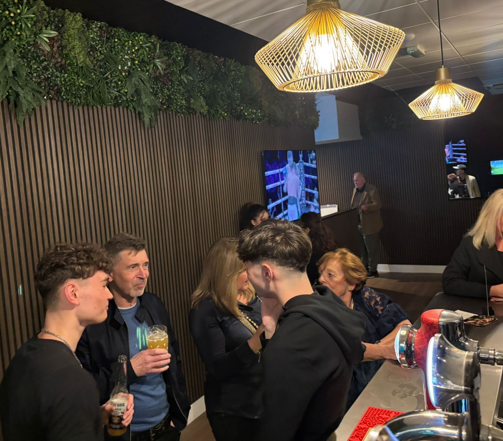 People gather at a bar, some drinking and conversing. The bar has wood paneling and modern pendant lights.
