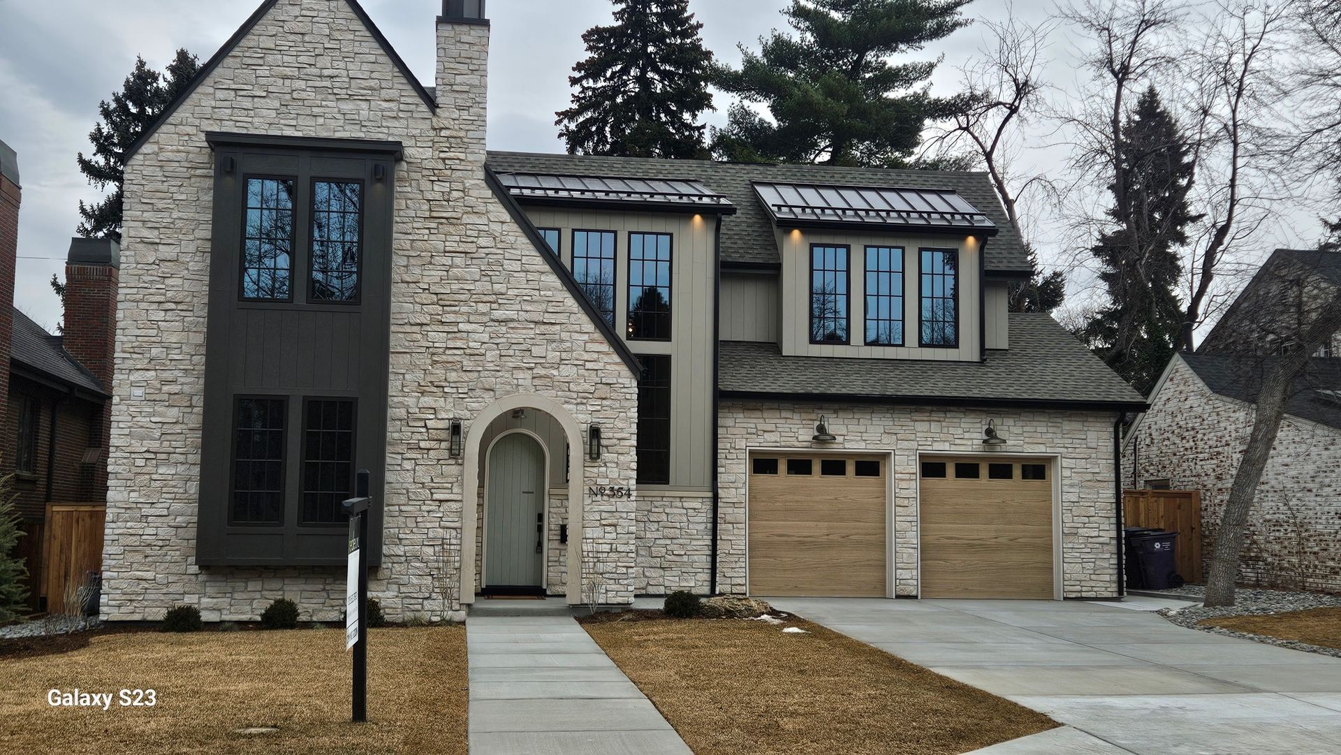 Stone-clad house with dark trim, arched door, and two-car garage. Driveway, walkway, and bare landscaping in front.