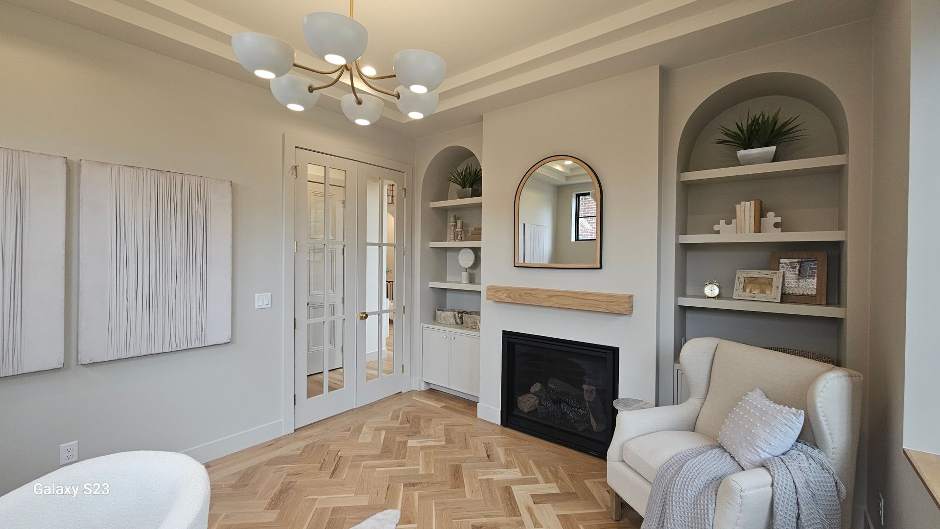 A bright home office with built-in arched shelves, fireplace, and light wood flooring. 