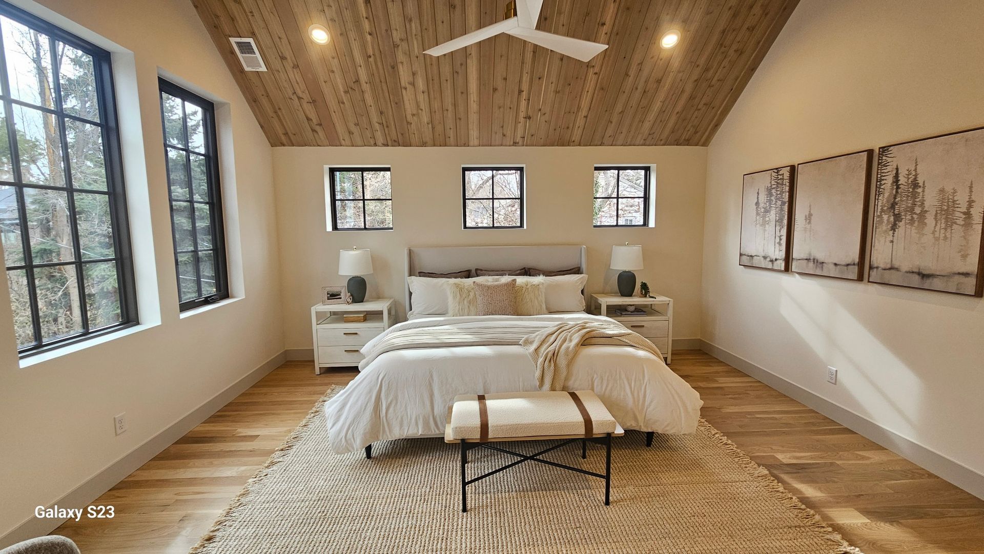 Bedroom with wood ceiling and floors, white walls, black-framed windows, bed, bench, rug, and artwork.